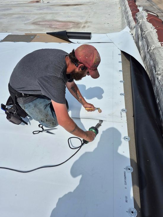 Man kneeling on a white roof welding a seam, wearing a hat, jeans, and gray shirt.