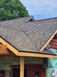 Newly shingled roof, brown shingles, wooden trim, red siding under construction, against a blue sky.