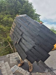 Roofer on a roof holding a large bundle of black asphalt shingles, trees in background.