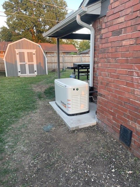 Generator next to a brick building. Small shed in the background, set on a grassy yard.