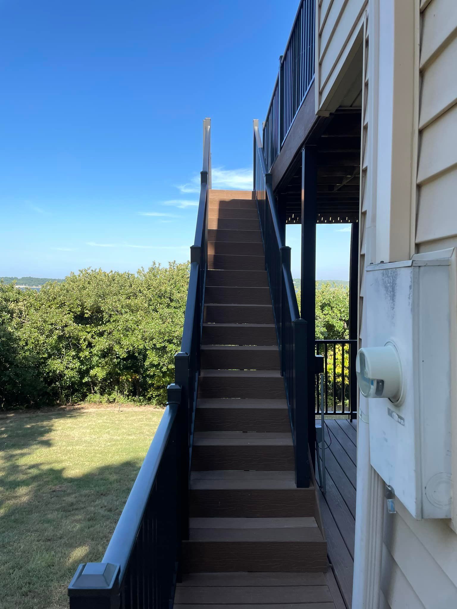 Wooden stairs leading up to a deck with black railings, against a backdrop of foliage and blue sky.