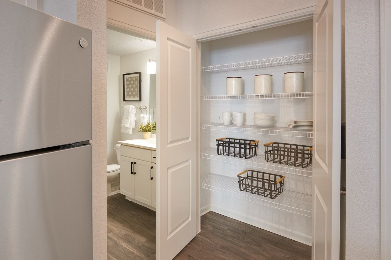 Open pantry closet with wire shelves and baskets beside a refrigerator in an apartment.