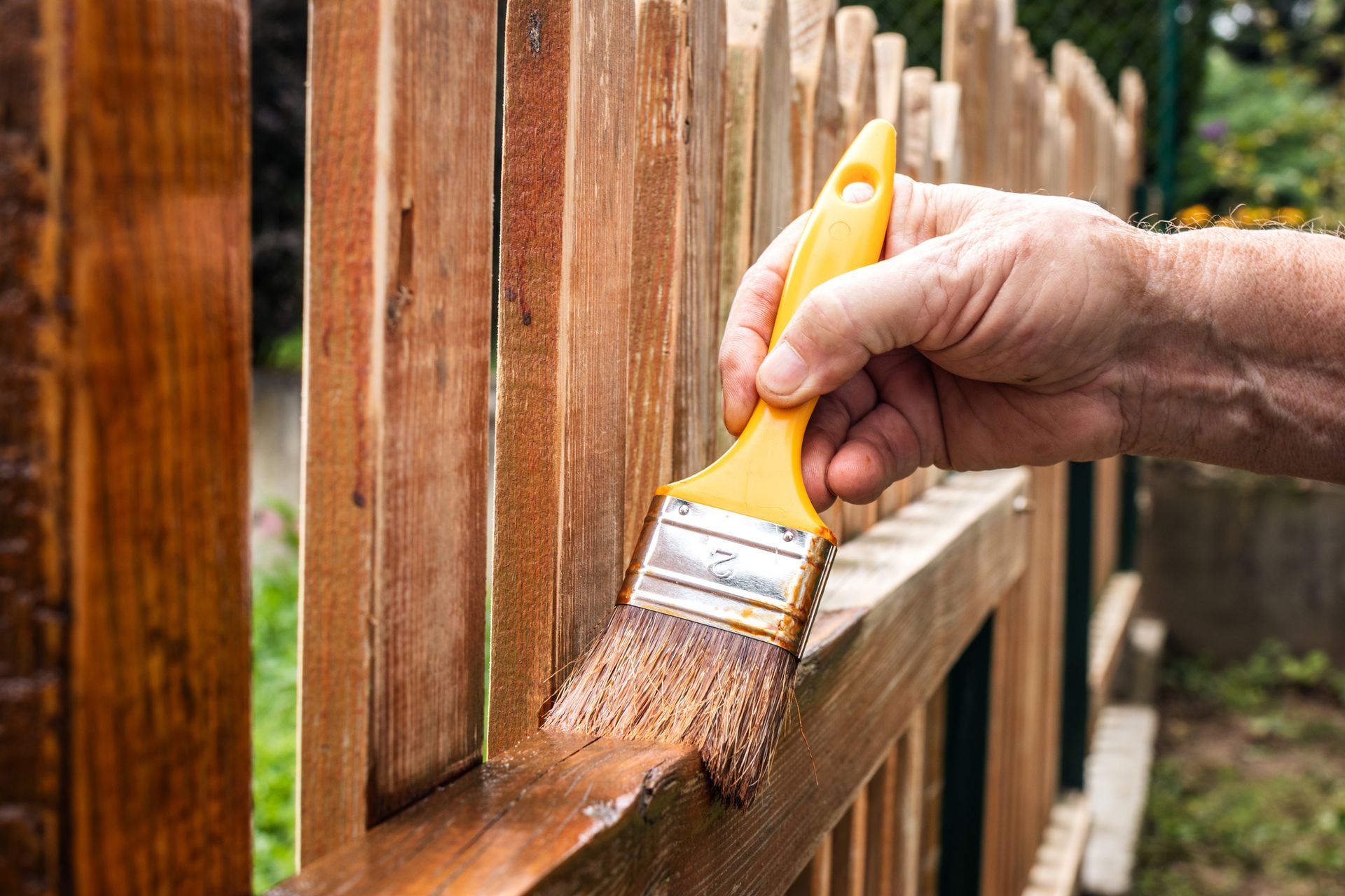 Person painting a wooden fence with a brush, applying a brown stain.