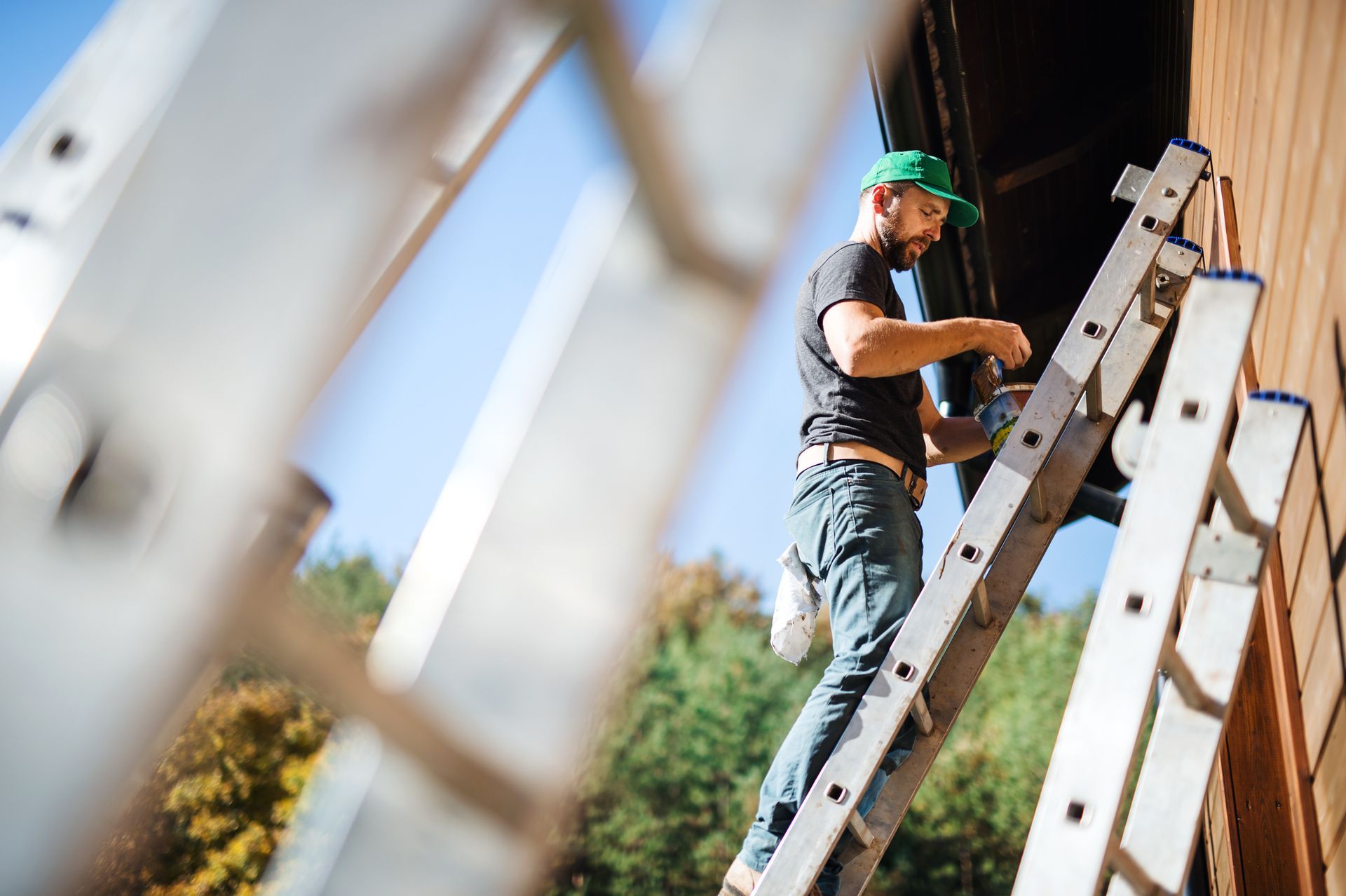 Man on a ladder, repairing a house gutter. He wears a green cap and jeans, outdoors on a sunny day.