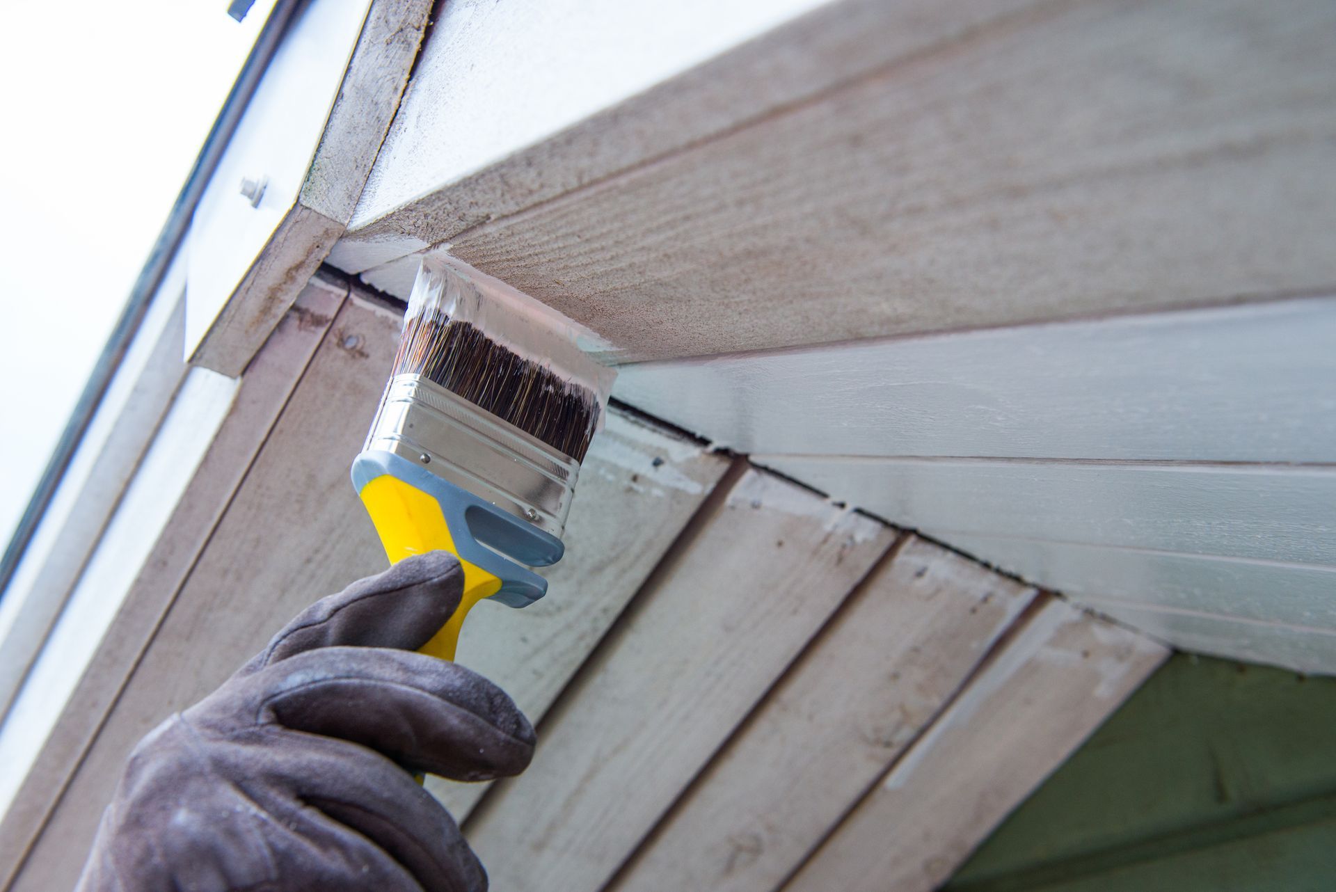 Person wearing work glove painting white trim on house's exterior with a paintbrush.