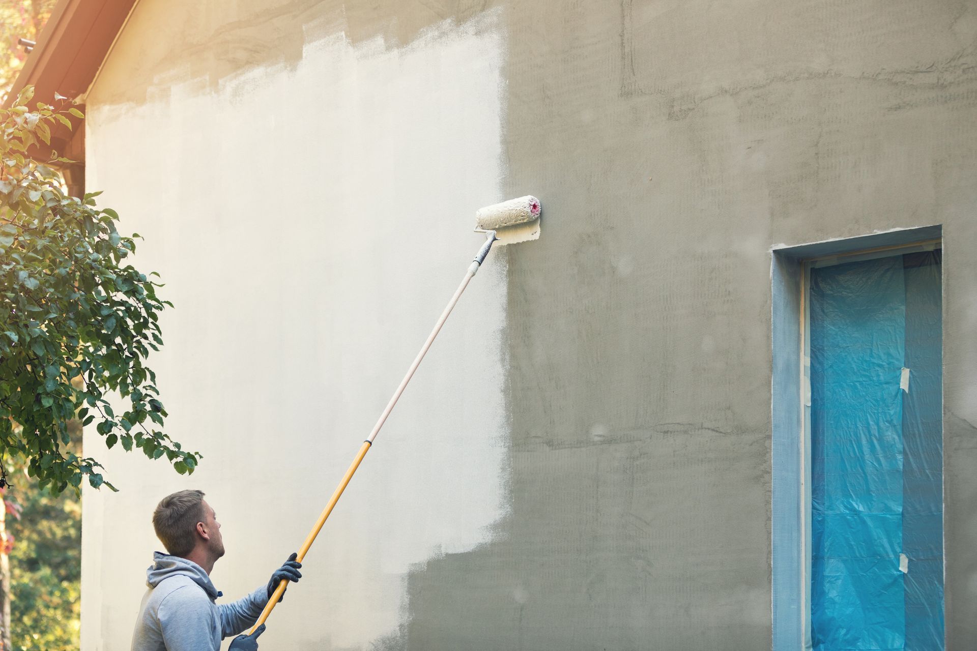 Man painting exterior wall with roller and extension pole; half wall painted light grey.