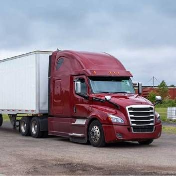 Red semi-truck with white trailer parked on a paved lot under a cloudy sky.