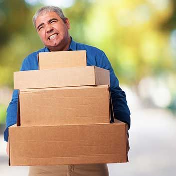 Man straining while carrying a stack of brown cardboard boxes outdoors.