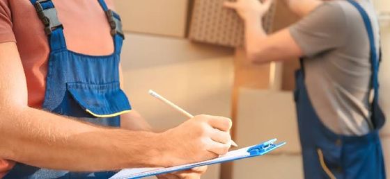 Two movers wearing overalls, one writing on a clipboard while another handles boxes.