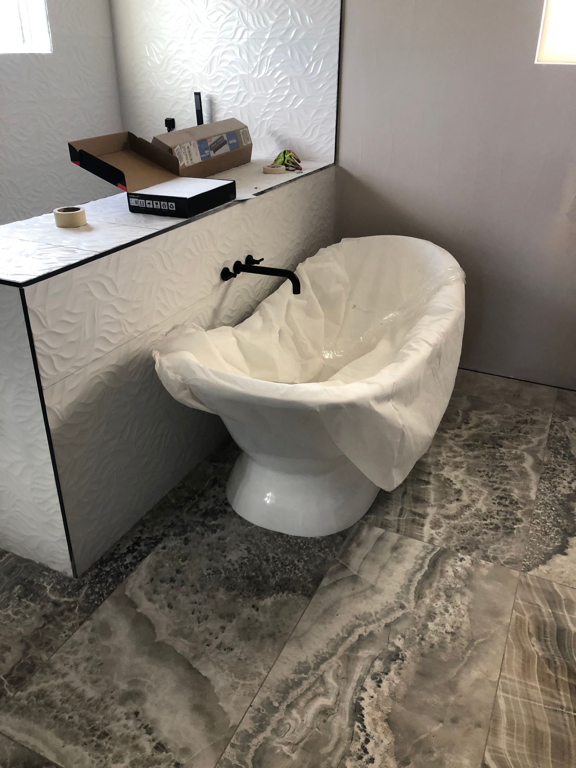 Freestanding white tub with protective cover in a bathroom. Gray marble-look floor and textured white wall detail.