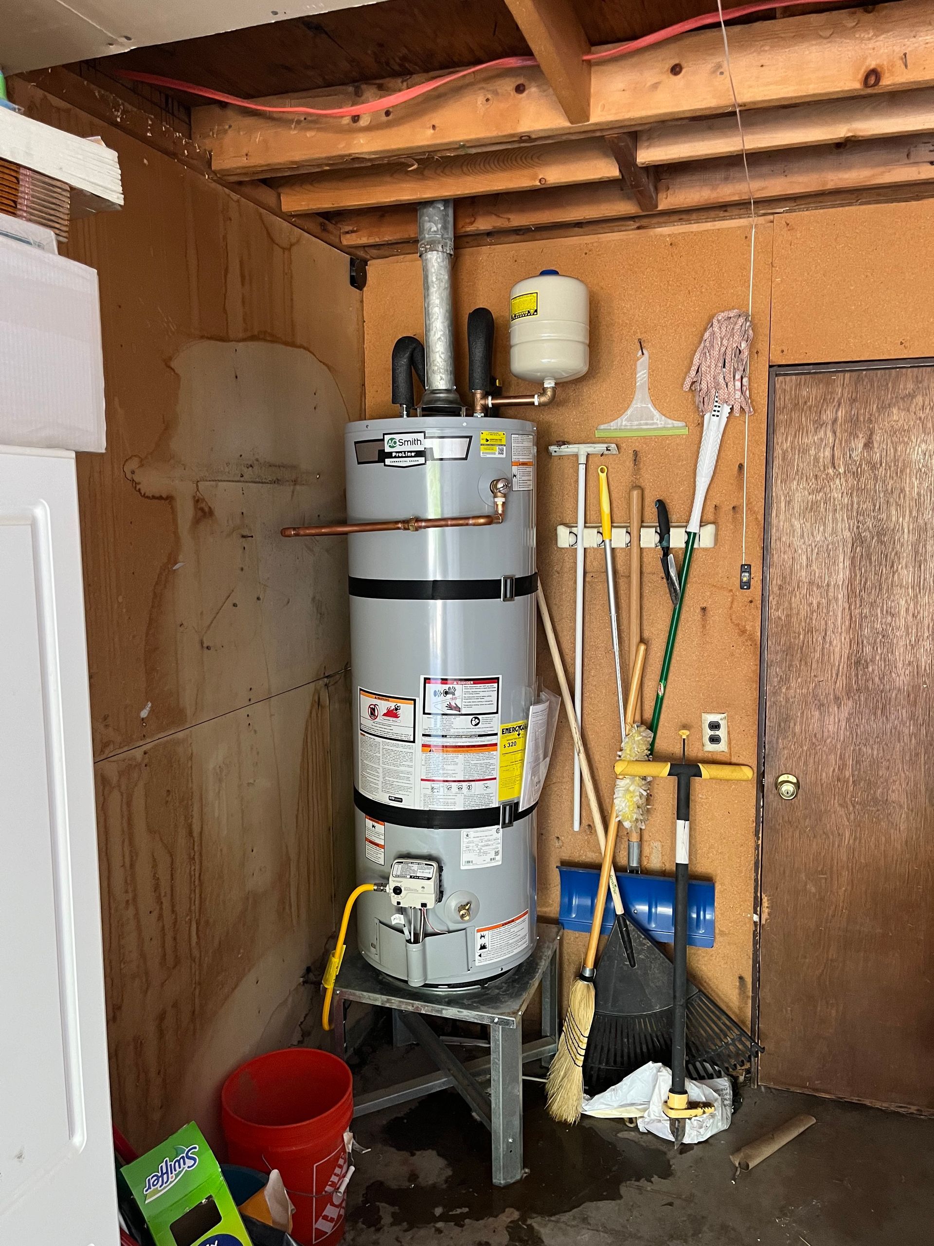 Water heater in a utility room with a pressure tank, various tools, and a red bucket.
