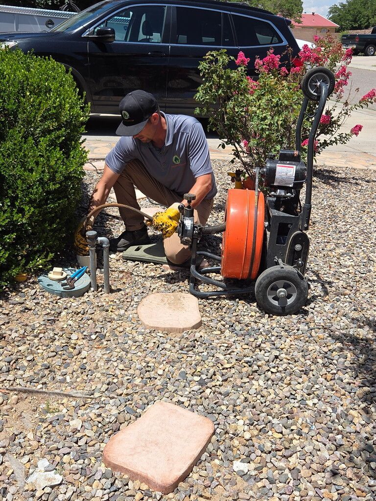 Plumber with a drain cleaning machine, working outdoors on a gravel area.