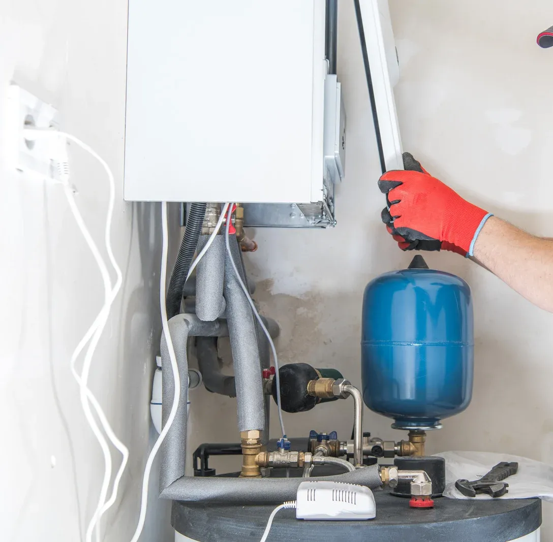Person in red gloves working on a heating system with a blue expansion tank and pipes in a room.