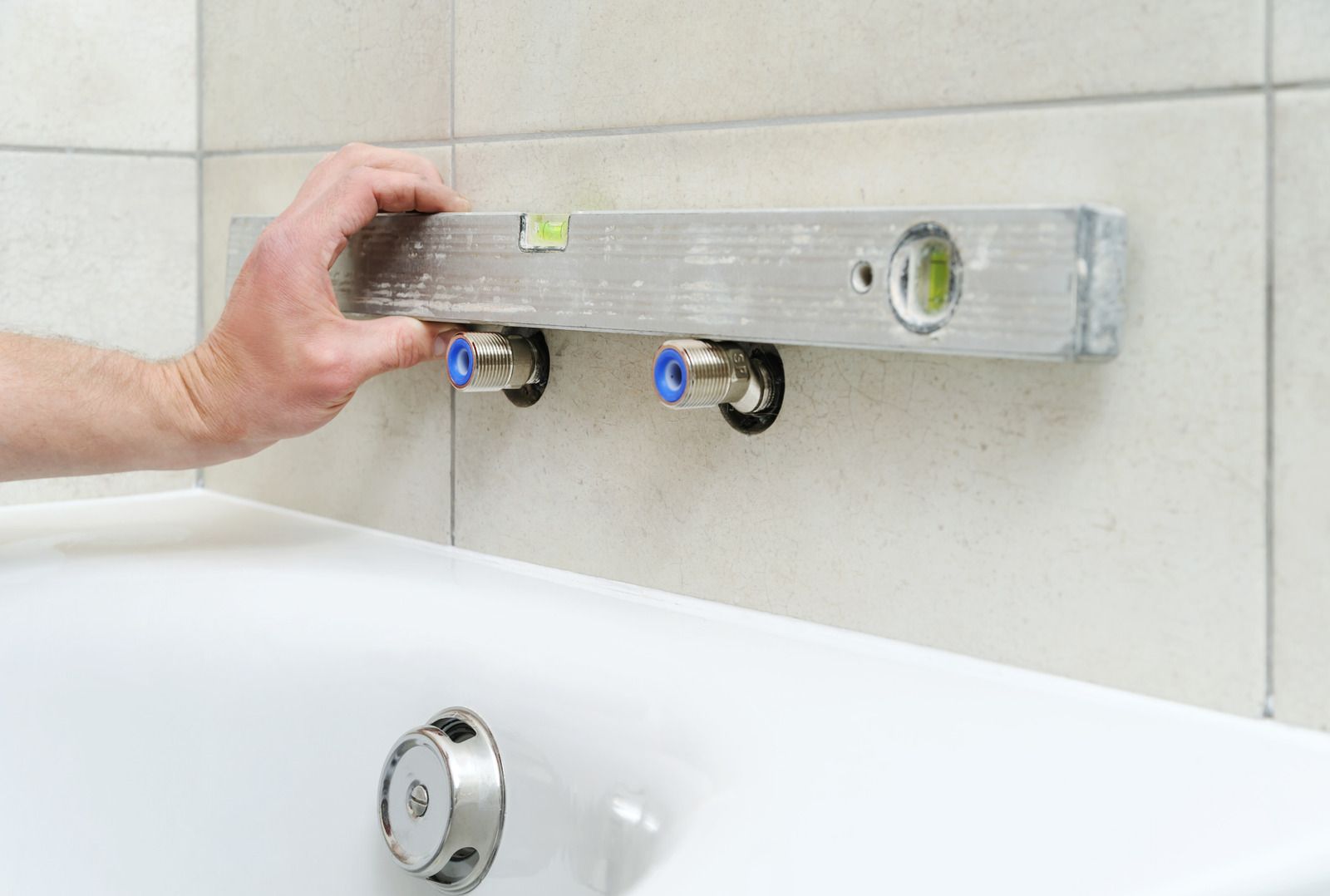 Person using a level to check the alignment of water pipe fittings on a tiled bathroom wall above a bathtub.