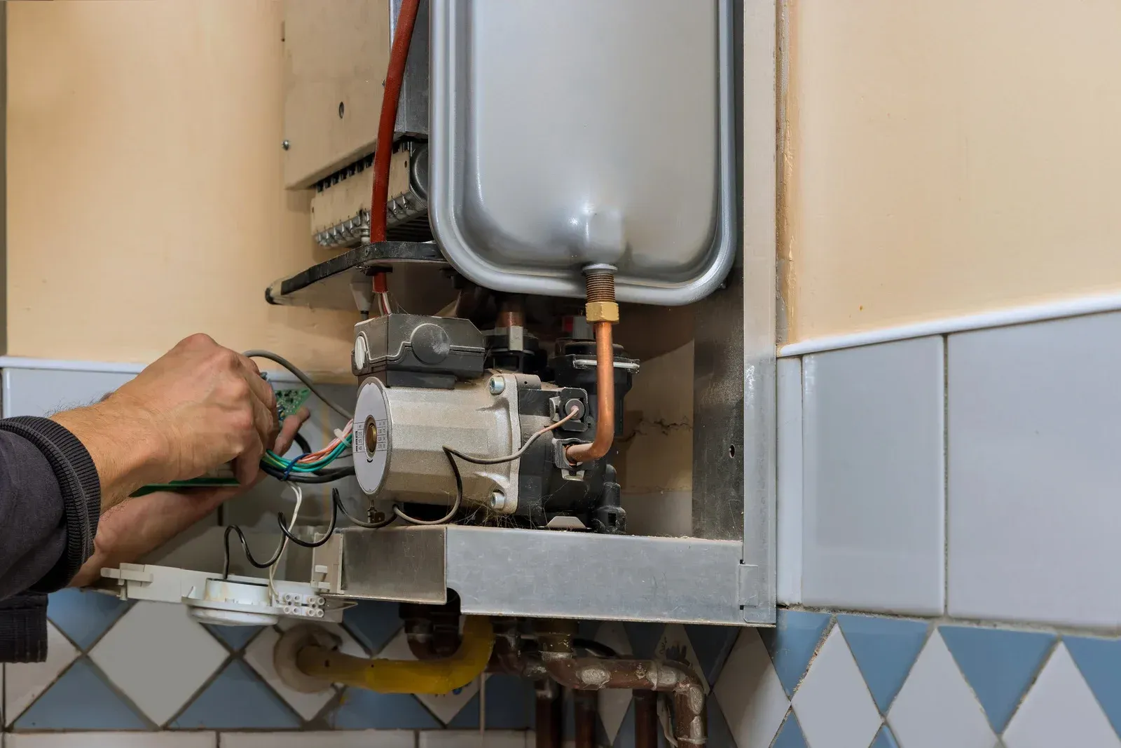 Person working on a water heater. Hands hold wires inside the open appliance on a tiled wall.