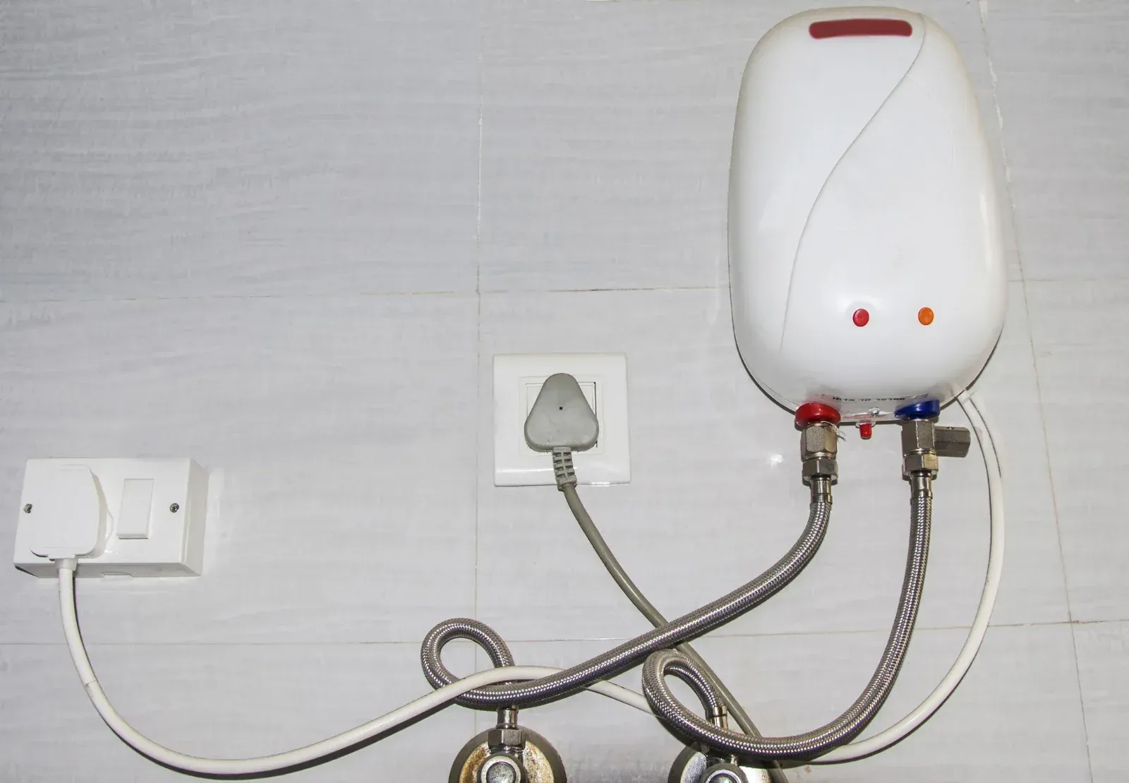 White electric water heater mounted on a wall with water pipes and a plug connected to a wall outlet.