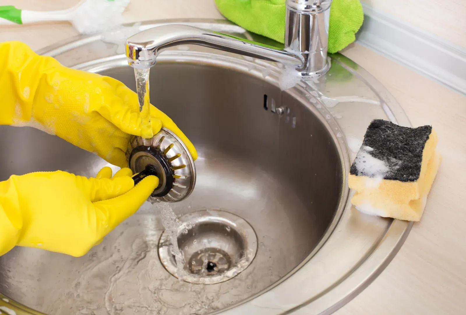 Hands in yellow gloves cleaning a kitchen sink drain strainer with running water.
