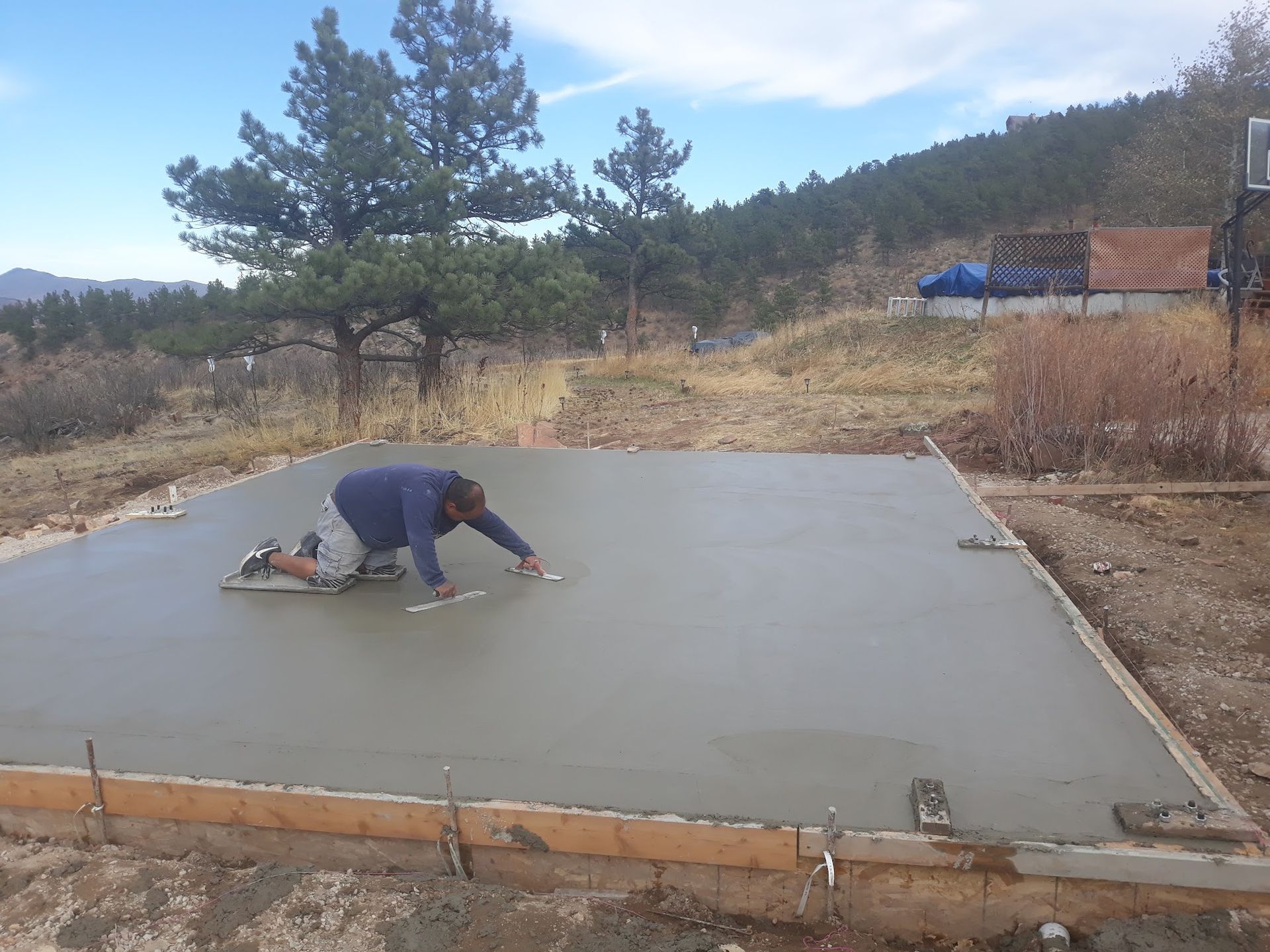 Person smoothing fresh concrete foundation outdoors. Blue sky, trees, and hillside in background.
