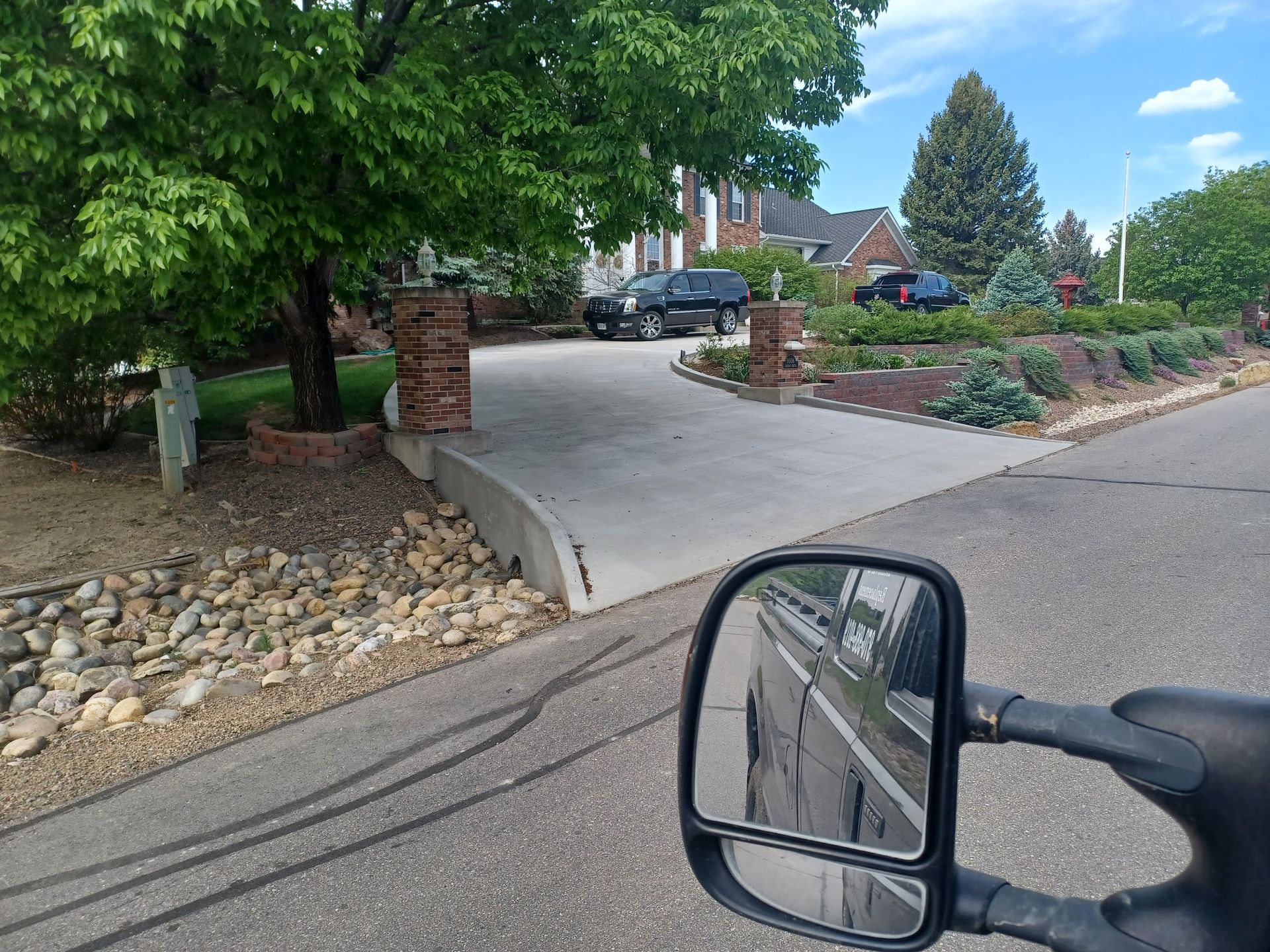 View from a vehicle's side mirror. Driveway to a large house with landscaping, trees, and parked vehicles.