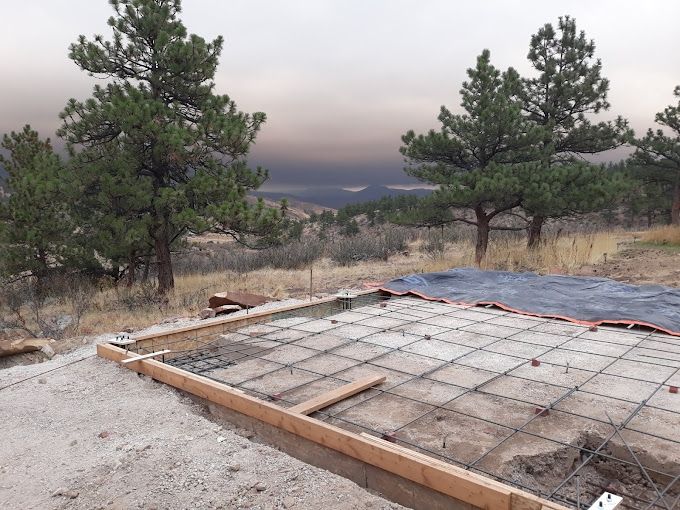 Concrete foundation under construction, framed by wood, with rebar grid. Trees and hazy sky in background.