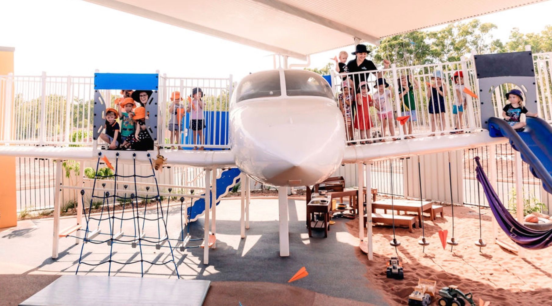 A group of children plays on an airplane-themed playground structure with a slide, climbing nets, and swings.
