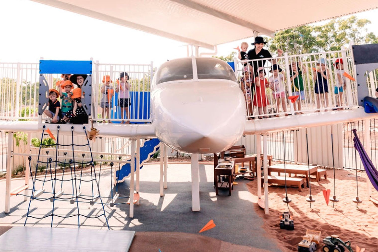 Children play on an airplane-themed playground structure with a cockpit, climbing net, and sand pit area under a canopy.
