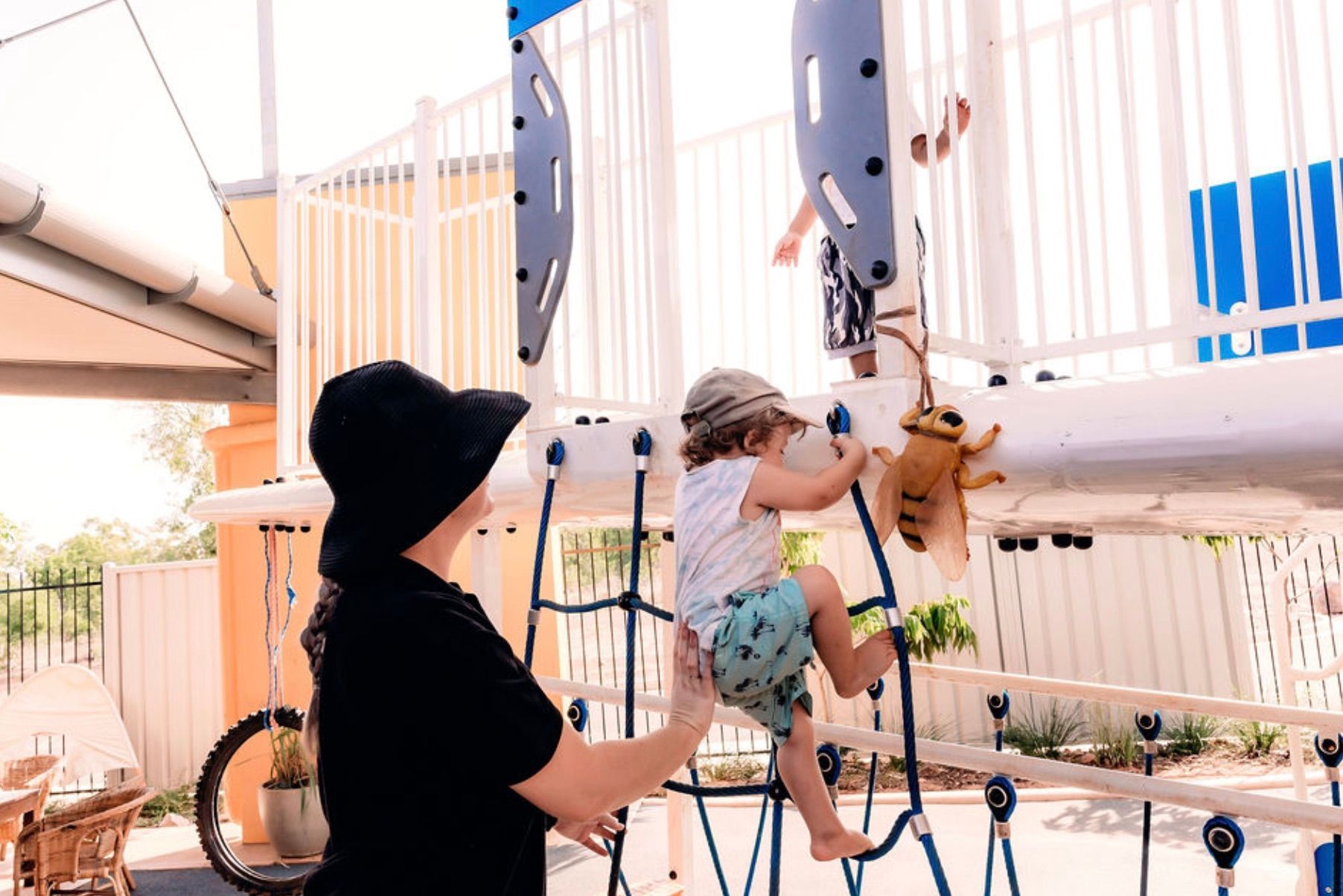 A person in a black hat assists a child climbing a rope playground structure.