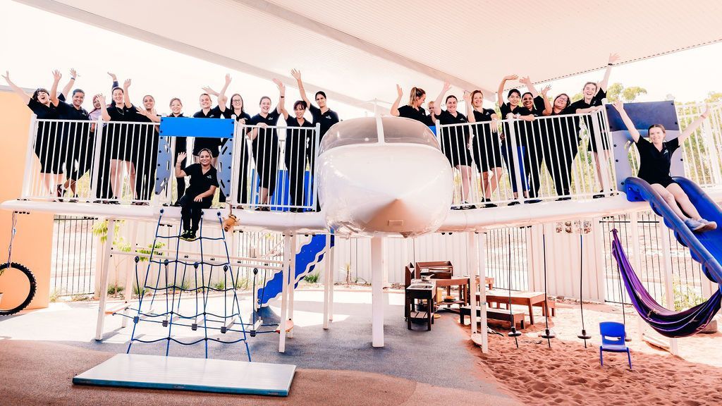 A group of people standing on and around an airplane-themed playground structure under a large canopy.