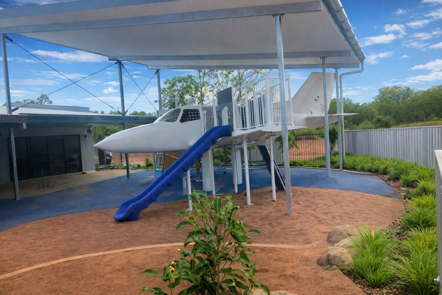 A white airplane-themed playground structure with a blue slide, positioned under a canopy in a sunny outdoor area.