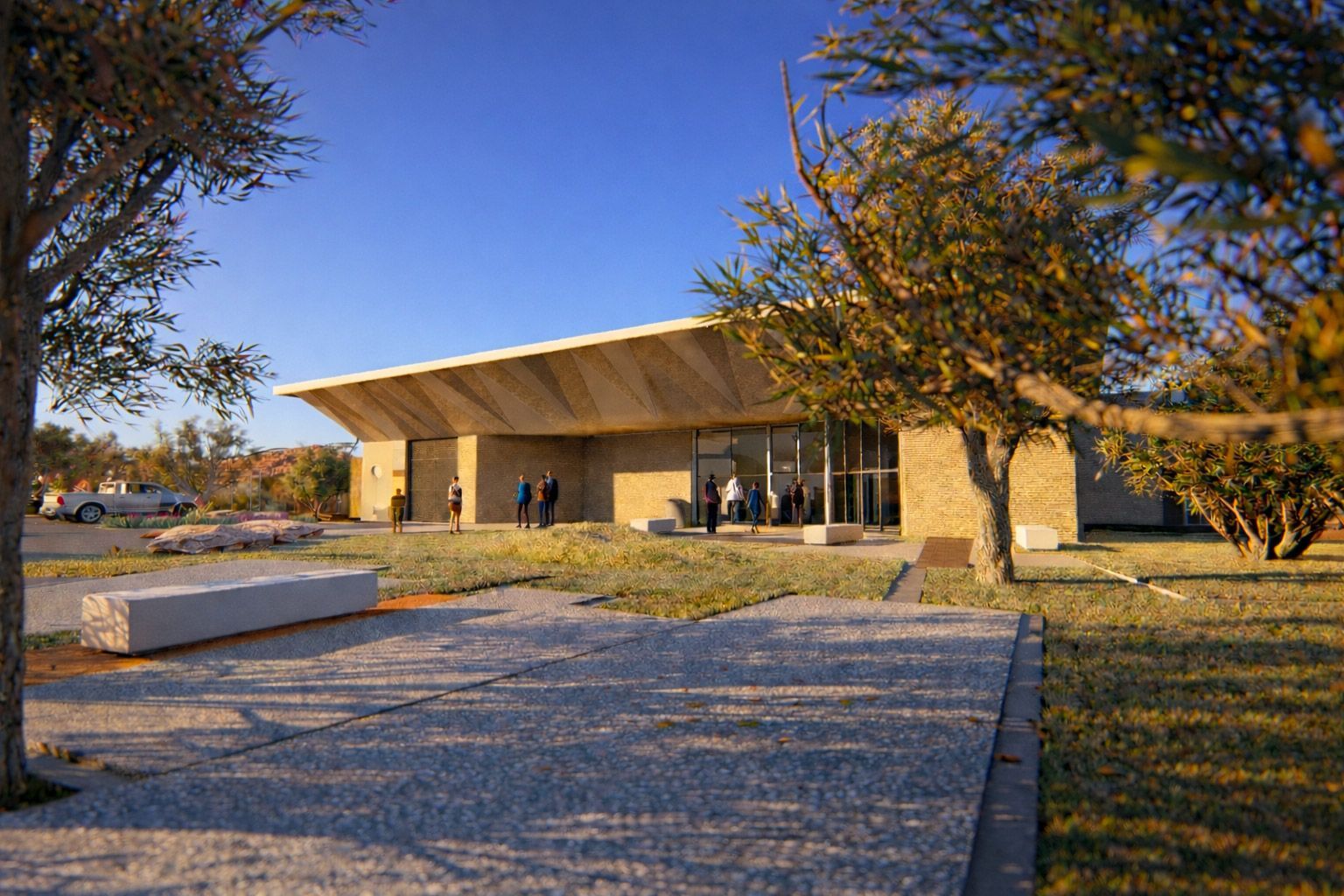 A modern, light-stone building with a wide, cantilevered roof sits in a sunny, arid landscape with trees and gravel paths.
