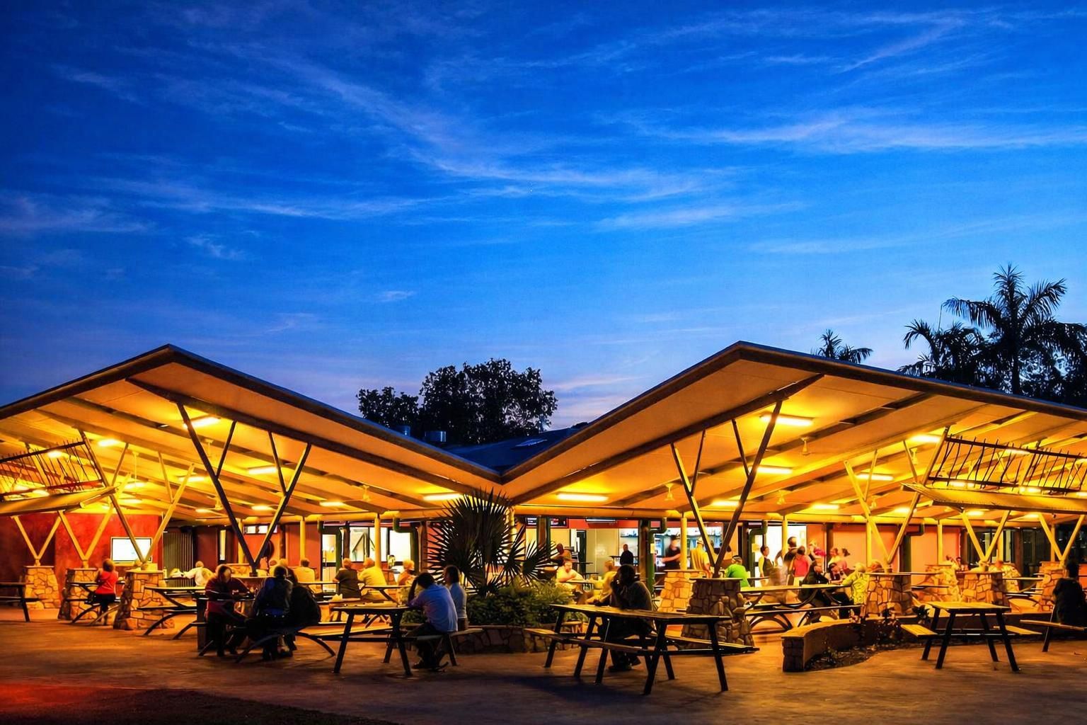 A modern playground under a large, petal-shaped shade structure in a wooded, sunlit park setting.