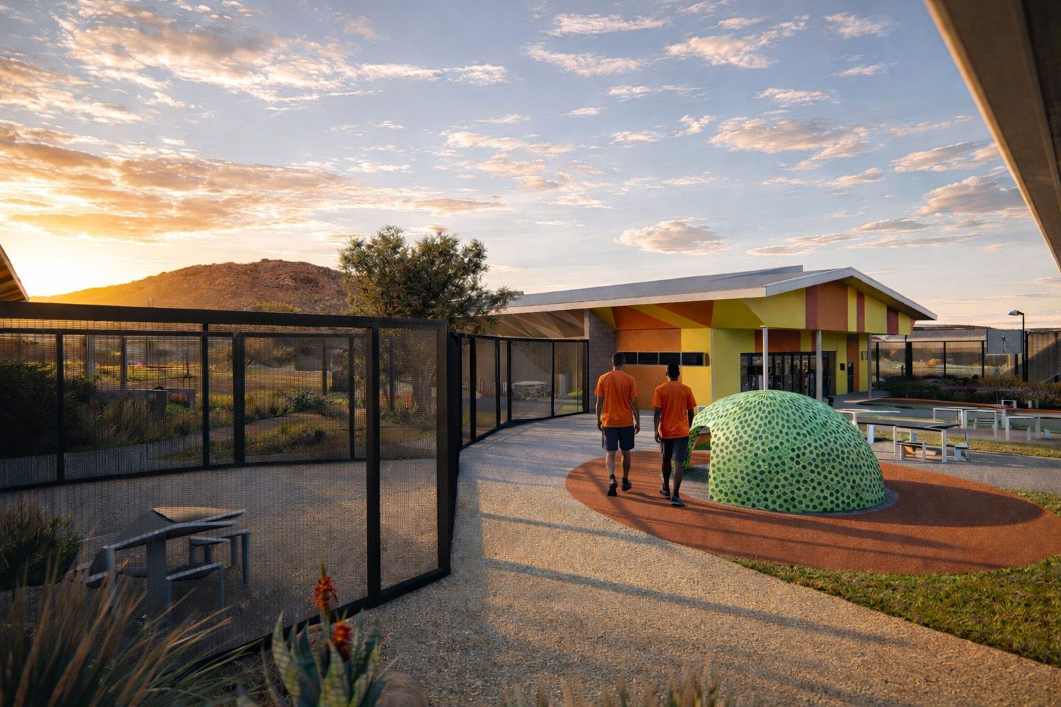 Two people walk on a gravel path toward a yellow-and-orange building at sunset, next to a green tiled dome structure.