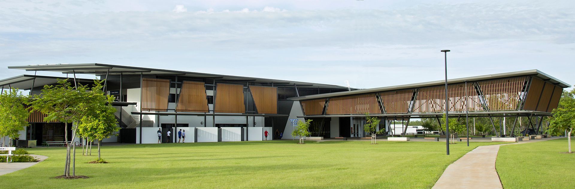 A modern, multi-level building with wood-slatted exterior panels, situated on a green lawn with a paved walkway.