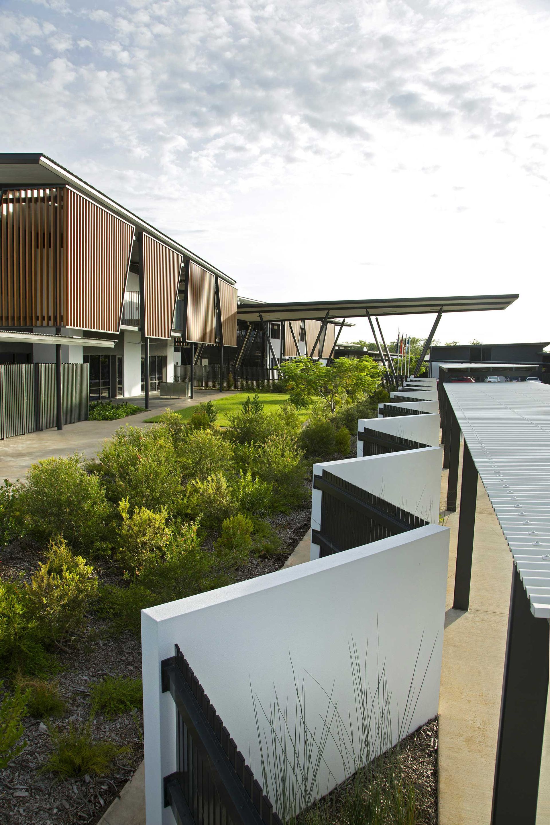 A zig-zagging white low wall borders a garden with green shrubs alongside a modern building with wooden exterior louvers.