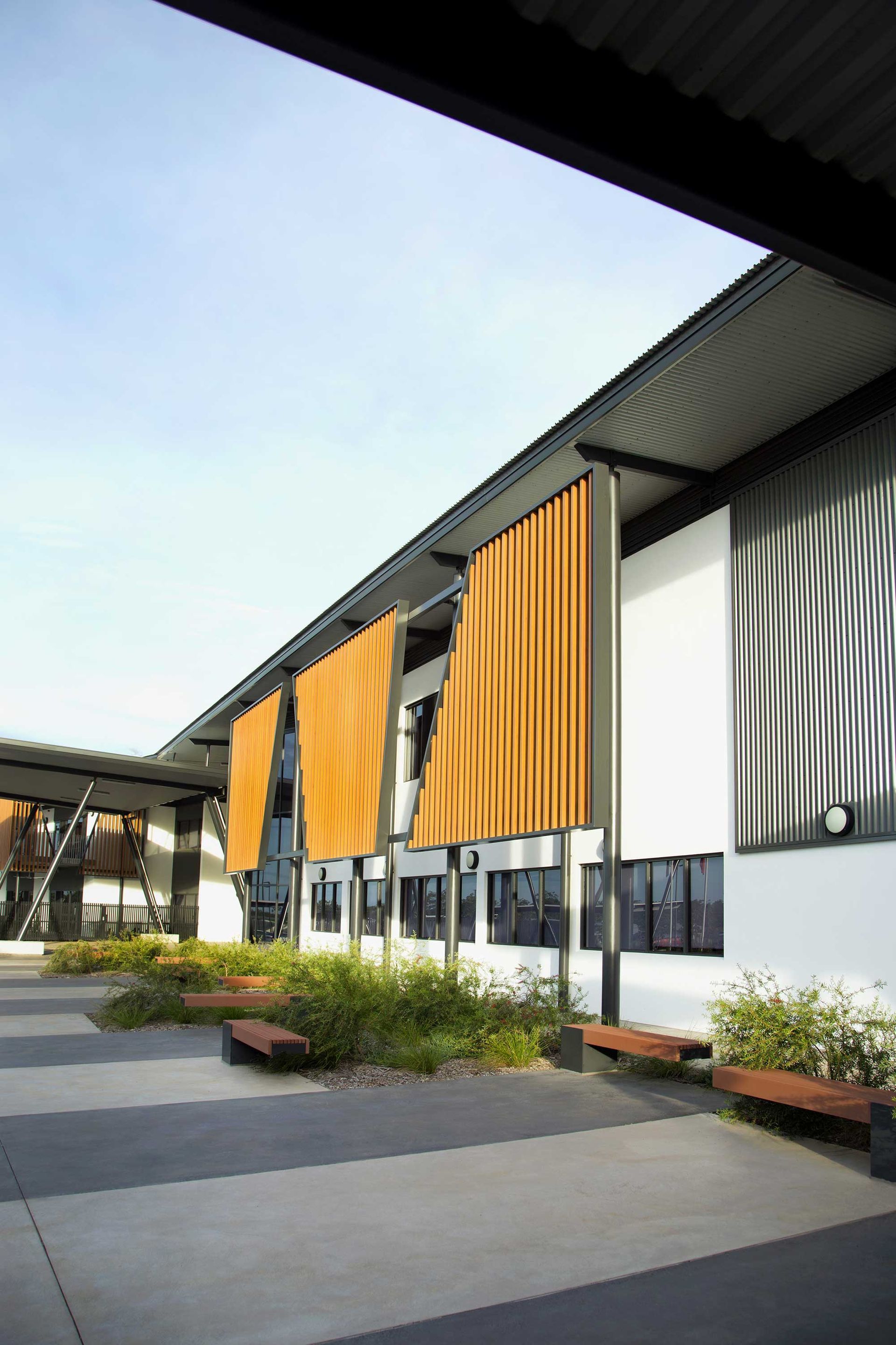 A modern school building courtyard with large, mustard-yellow fabric sunshades, a concrete patio, and wooden benches.