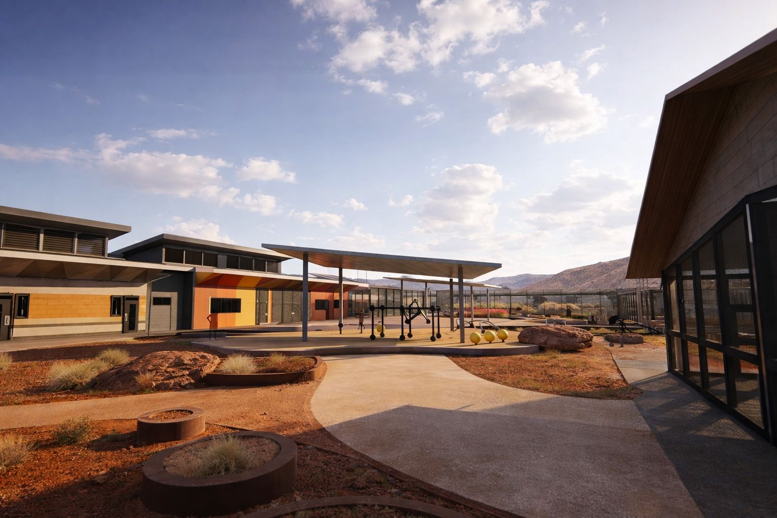 A sunlit courtyard in a desert landscape with multi-colored buildings, a central shaded structure, and rocky landscaping.