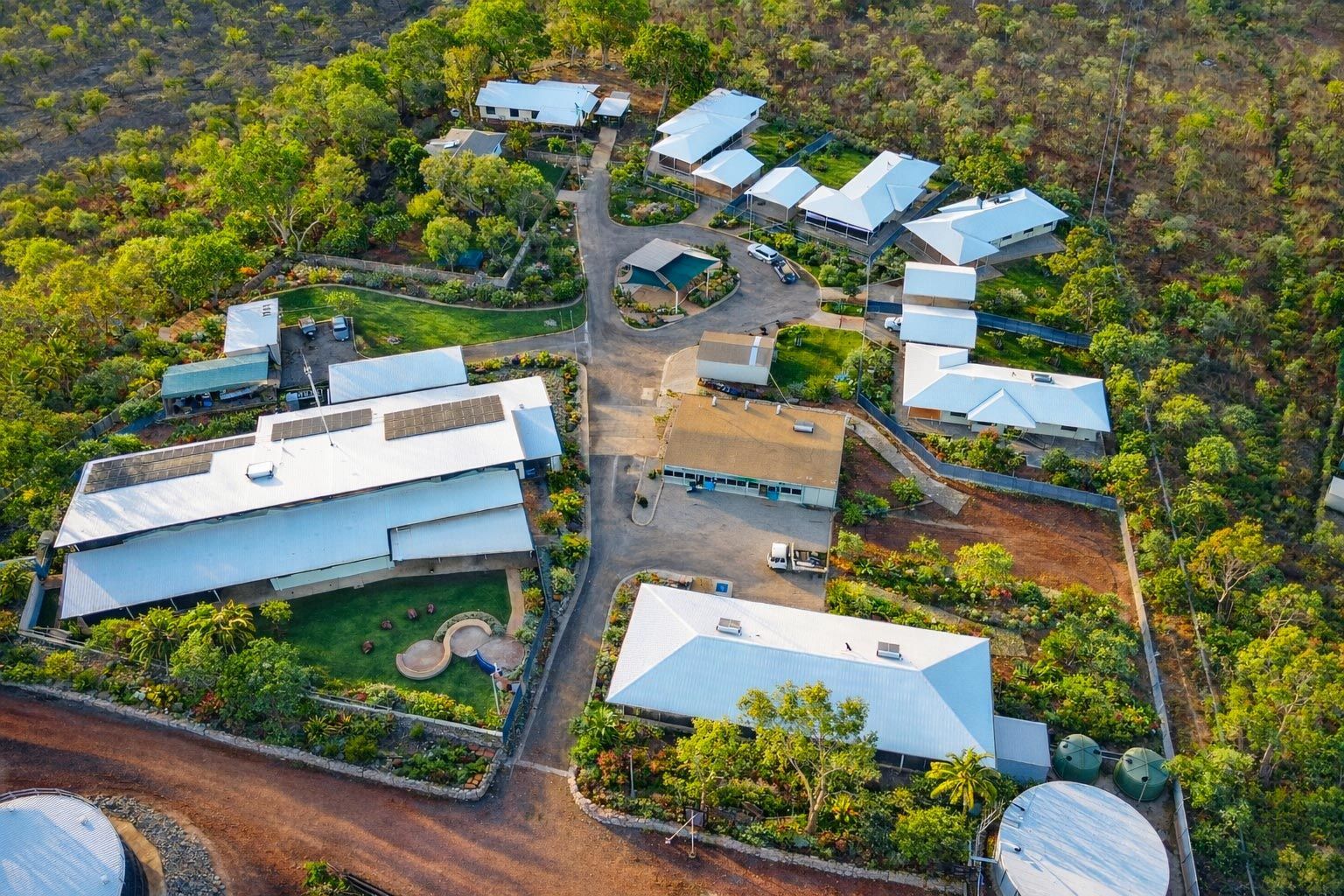 An aerial view of a remote lodge with white-roofed buildings, green landscaping, and gravel paths surrounded by bushland.