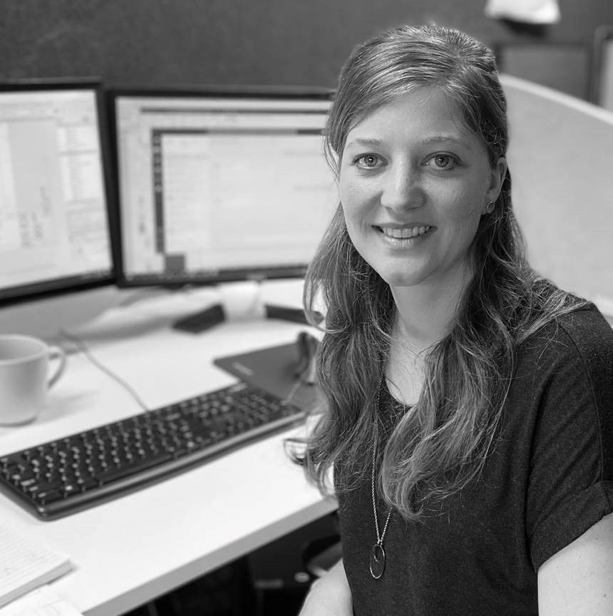 A smiling person sits at an office desk with two computer monitors, a keyboard, and a mug.