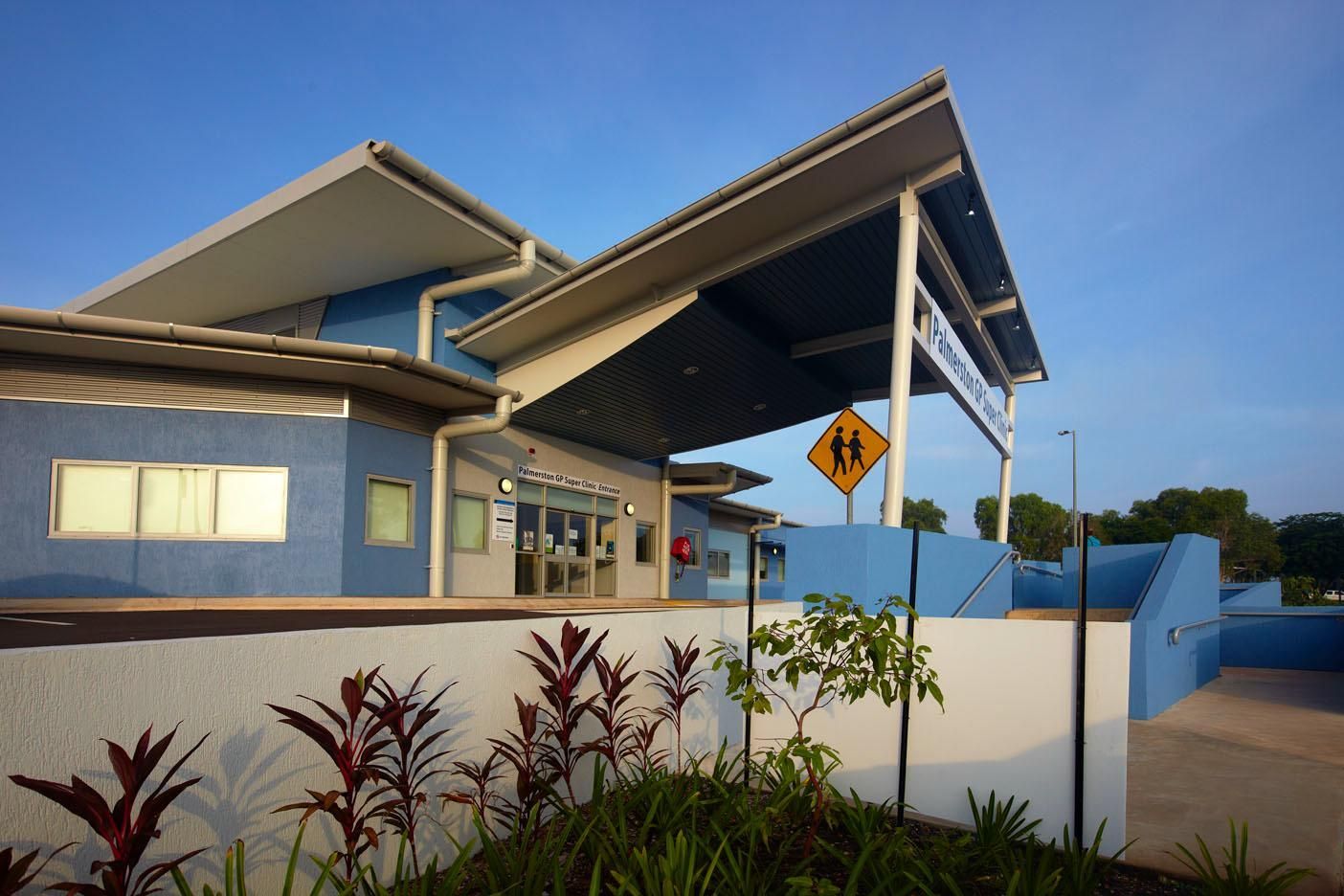 A modern blue community building with a large covered entrance, a pedestrian crossing sign, and landscaping out front.