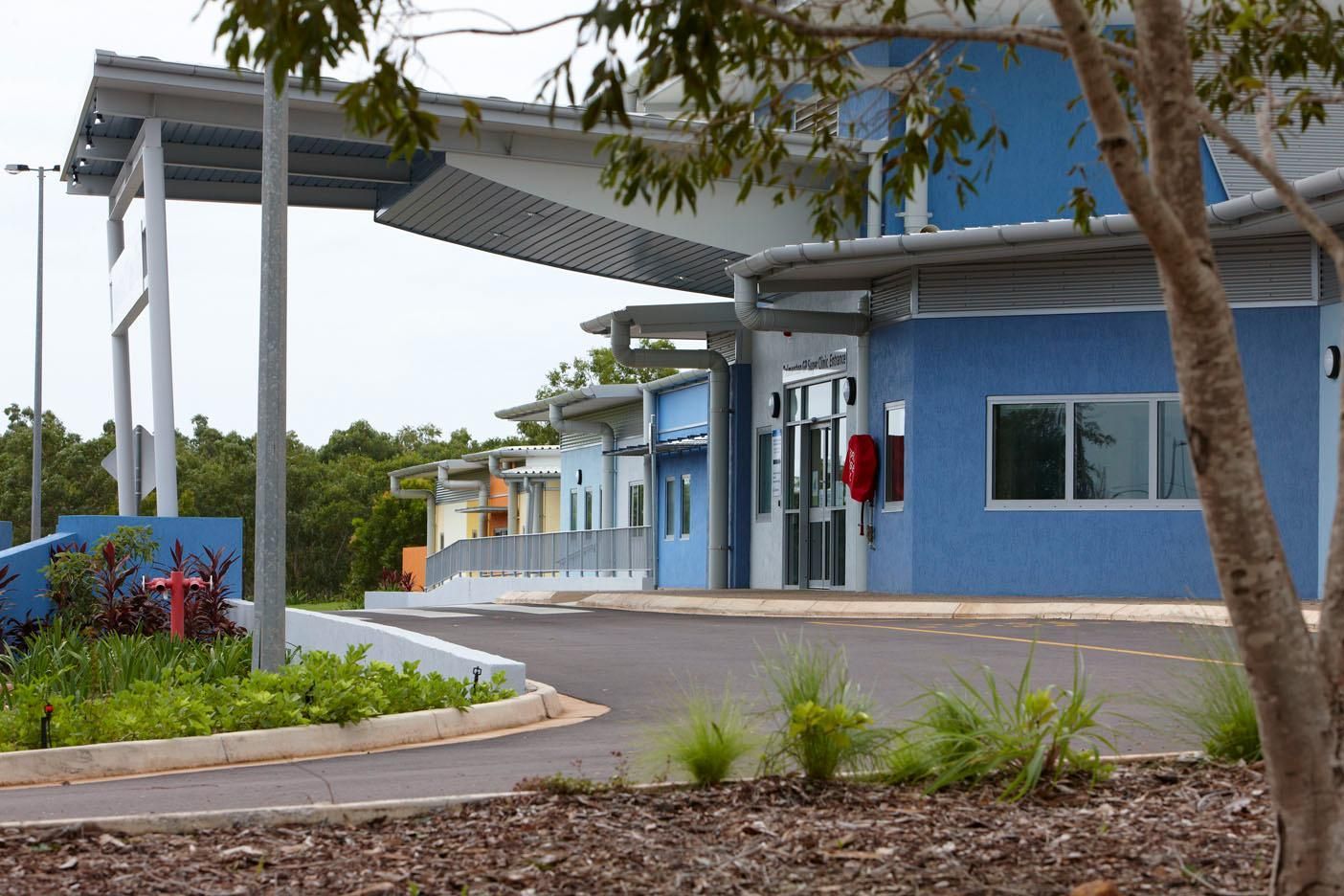 The blue facade of a modern, single-story building with a covered walkway and paved driveway surrounded by landscaping.