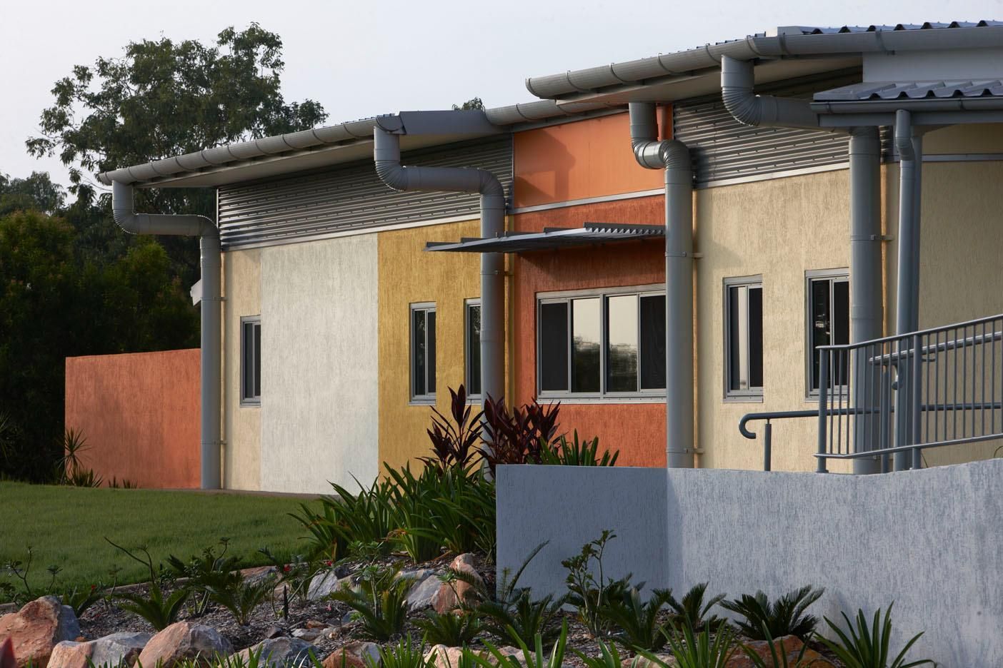 A modern, one-story building with a multi-colored stucco facade, metal roof, and an accessible ramp near a garden.