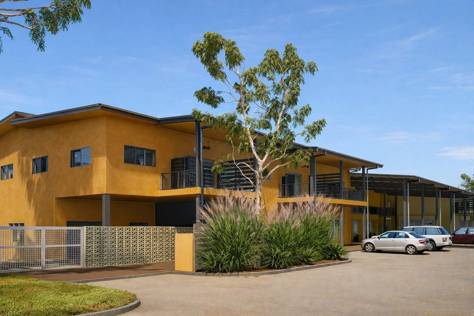 A modern two-story yellow building with a metal roof and a gravel parking lot with parked cars under a sunny blue sky.