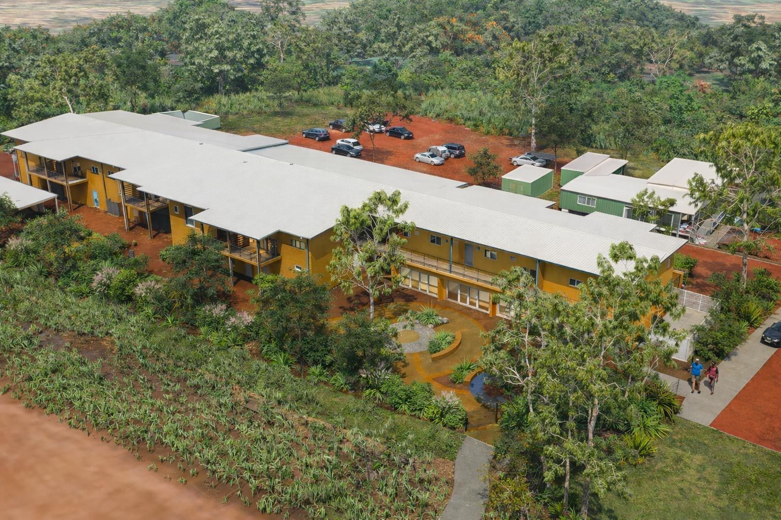 A long, yellow, one-story building with a white roof nestled in a lush, green landscape with a red dirt parking lot.