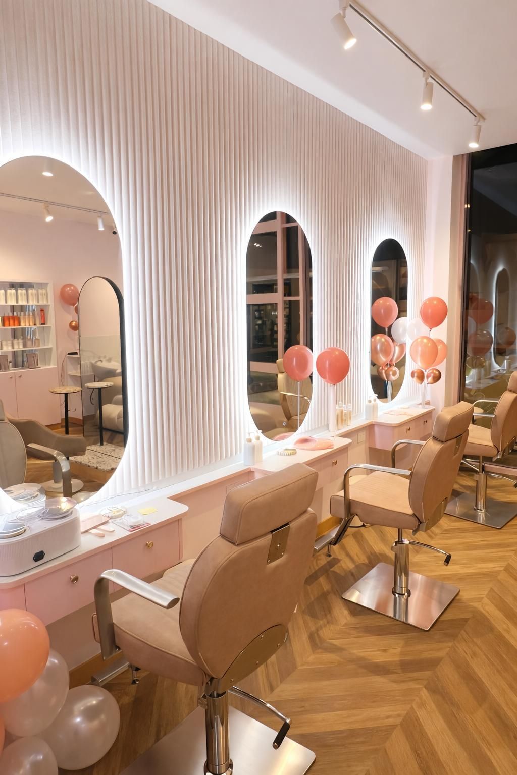 A bright salon with three arched vanity mirrors, pastel pink styling chairs, wooden floors, and decorative peach balloons.
