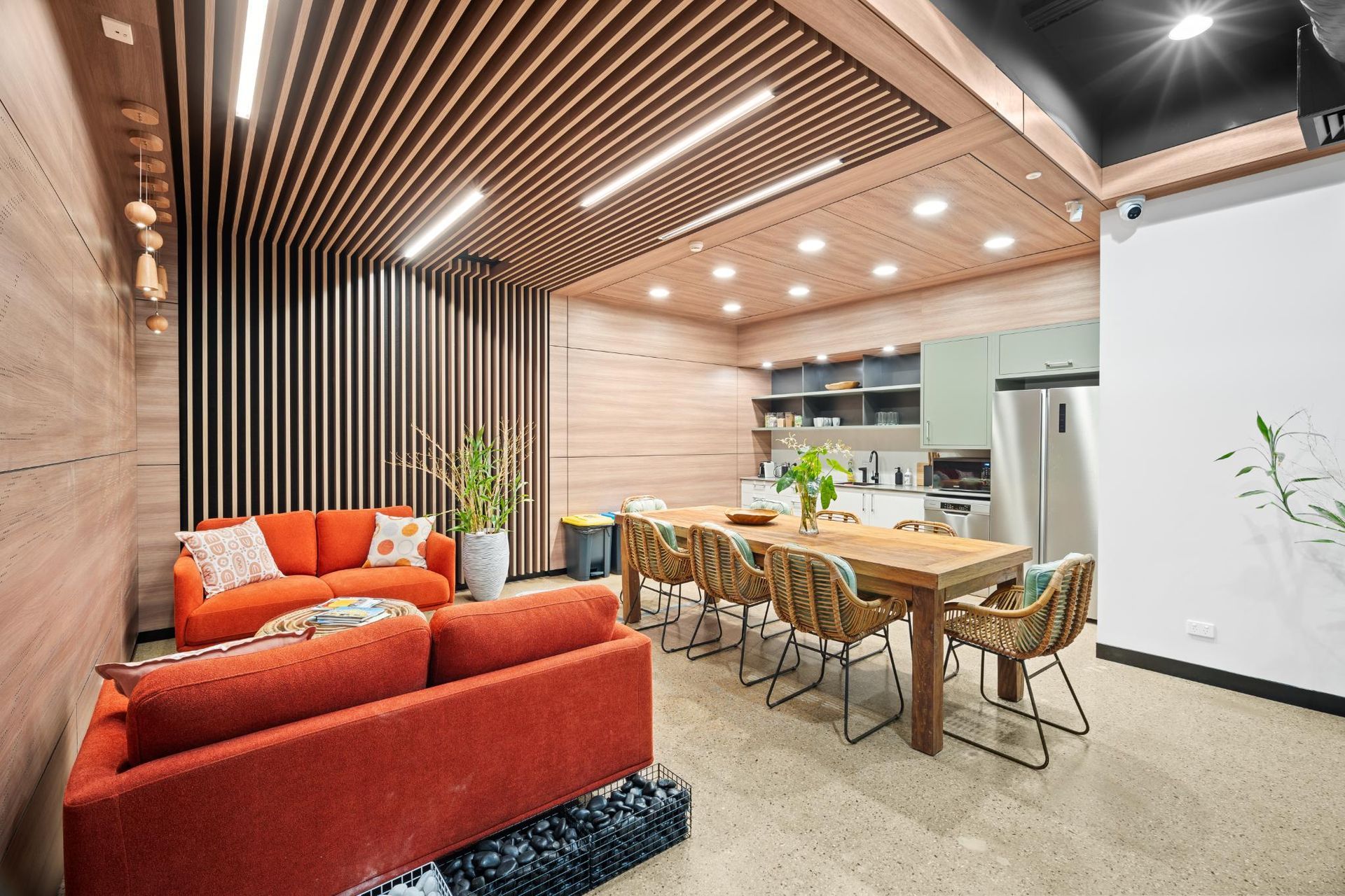 A modern communal kitchen and lounge with orange sofas, a wooden dining table, and slatted wood ceilings.