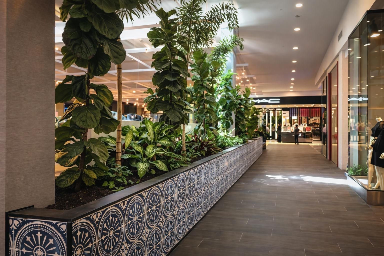 Indoor mall corridor featuring a long, tiled planter box with tall green plants and trees beside storefronts.