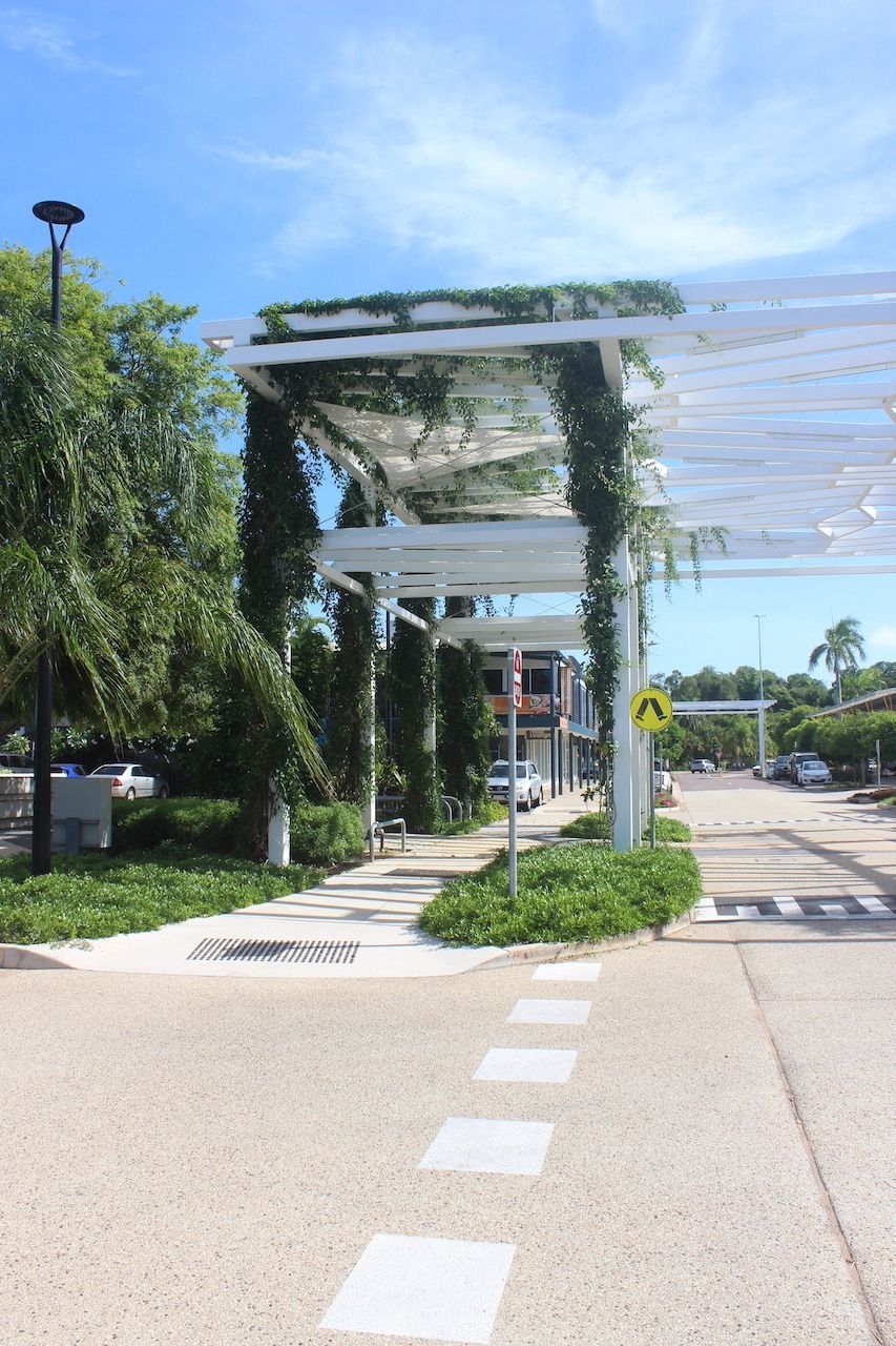 A white wooden pergola covered in climbing plants shades a pedestrian walkway next to a road with a yellow warning sign.