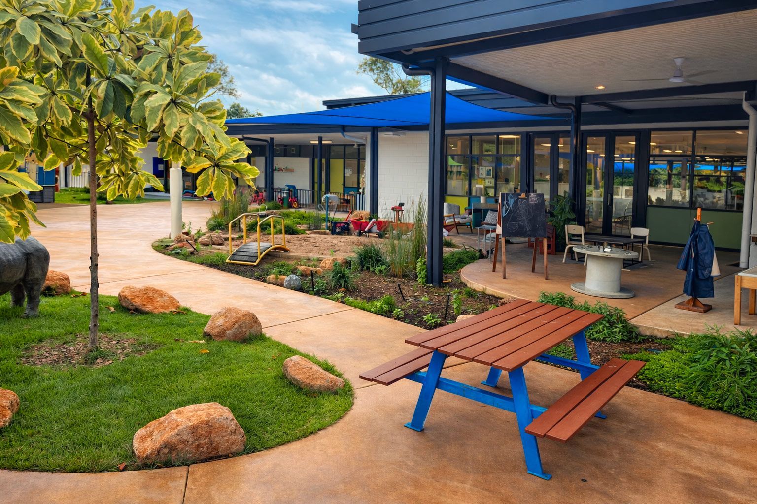A sunny outdoor preschool courtyard featuring a blue picnic table, a landscaped play area with rocks, and a covered patio.