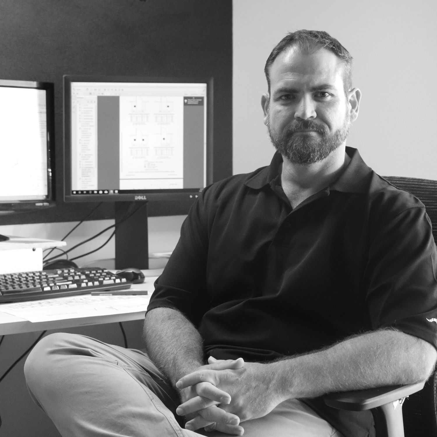 A person wearing a patterned button-down shirt smiles while working with a pen on technical drawings at a glass desk.