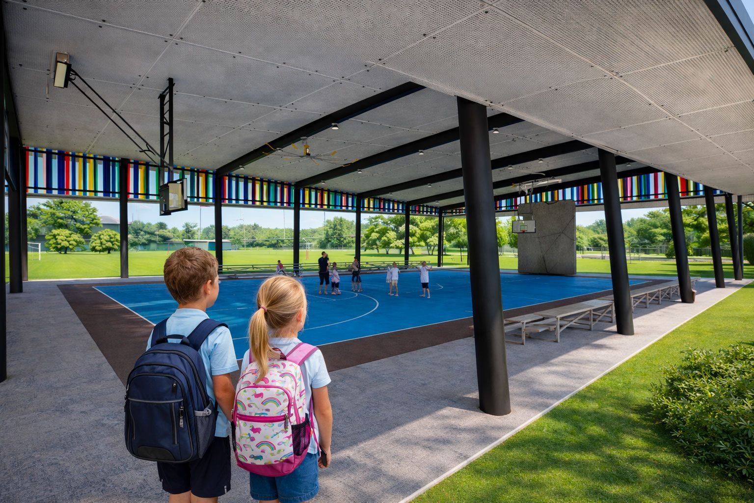 Two children with backpacks stand on a patio looking out at an outdoor sports court with a blue surface under a roof.